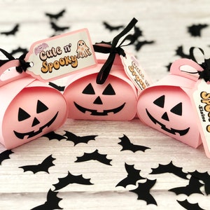 Puede incluir: Tres cajas de regalo rosas con tem&aacute;tica de Halloween con forma de calabaza, cada una con una cara de jack-o'-lantern negra. Cinta negra y etiquetas que dicen "Cute n' Spooky" con un dise&ntilde;o de fantasma y murci&eacute;lago. Recortes de murci&eacute;lagos negros esparcidos.