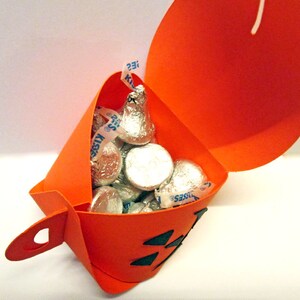 Puede incluir: Un recipiente naranja de papel con forma de calabaza lleno de caramelos de chocolate envueltos en papel de aluminio. El recipiente tiene una cara de Jack-o'-lantern recortada y un asa. Los caramelos son Hershey's Kisses.
