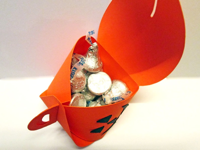 Puede incluir: Un recipiente naranja de papel con forma de calabaza lleno de caramelos de chocolate envueltos en papel de aluminio. El recipiente tiene una cara de Jack-o'-lantern recortada y un asa. Los caramelos son Hershey's Kisses.
