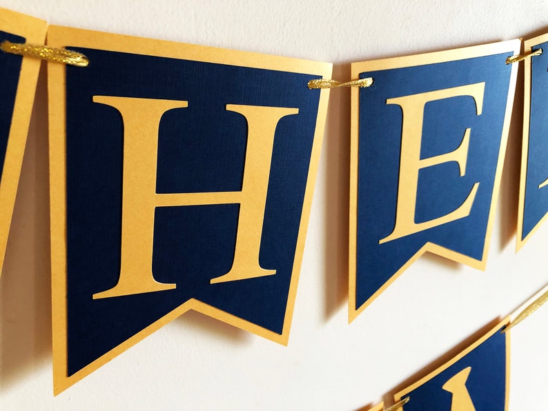 May include: A navy blue banner with gold lettering spelling out a word. The banner is made of individual pennant-shaped flags with gold trim and strung together with gold string. The letters are large and easy to read.