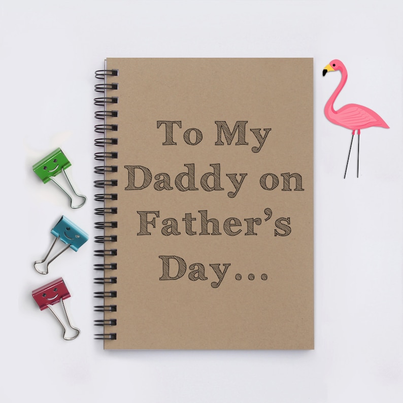 May include: A spiral-bound notebook with a kraft paper cover, featuring the text "To My Daddy on Father's Day...". Three binder clips in green, blue, and red with smiling faces are to the left. A pink flamingo decoration is on the right.