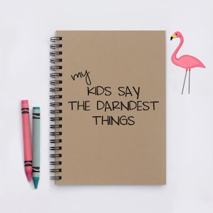 May include: A brown notebook with a spiral binding and the text "my kids say the darnedest things" written on it. There are two crayons, one pink and one blue, next to the notebook. A pink flamingo cutout is on the right side of the image.