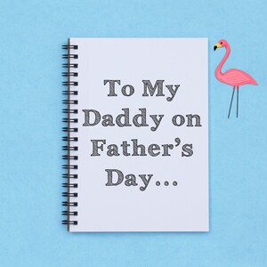 May include: A white spiral-bound notebook with the text "To My Daddy on Father's Day..." in black. A pink flamingo decoration is on the right side of the notebook, set against a light blue background.