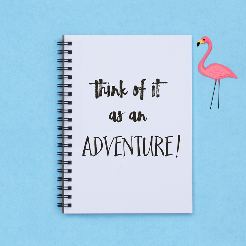 May include: A white notebook with a black spiral binding and the text "Think of it as an adventure!" written in black marker. A pink flamingo figurine is on the right side of the image.