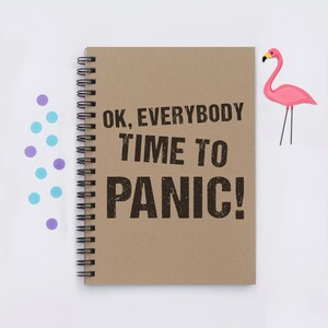 May include: A brown notebook with a spiral binding and the text "OK, EVERYBODY TIME TO PANIC!" printed in black. A pink flamingo figurine is on the right side of the image.