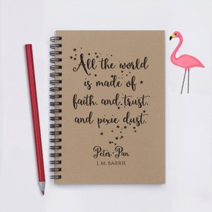 May include: A brown notebook with a spiral binding features a quote from Peter Pan: "All the world is made of faith, and trust, and pixie dust." The quote is printed in black with small stars surrounding the text. The notebook is on a white surface with a pink flamingo figurine to the right.