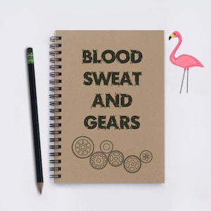 May include: A brown notebook with a spiral binding. The cover has the text "BLOOD SWEAT AND GEARS" in black outlined letters. There are several black and white gear illustrations below the text.