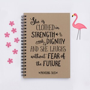 May include: A brown notebook with a spiral binding features a quote about strength and dignity. The quote reads "She is clothed in strength and dignity and she laughs without fear of the future." The quote is attributed to Proverbs 31:25.