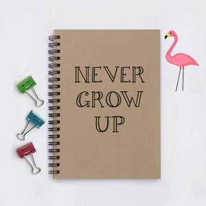 May include: A brown notebook with a spiral binding and the text "NEVER GROW UP" printed in black. There are three colorful paper clips and a pink flamingo cutout on a white background.