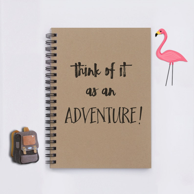 May include: A brown notebook with a spiral binding and the text "Think of it as an adventure!" written in black. A pink flamingo cutout is on the right side of the notebook. A brown backpack is on the left side of the notebook.