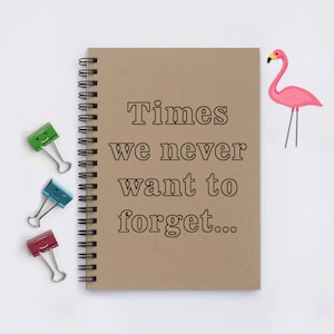 May include: A brown notebook with a spiral binding and the text "Times we never want to forget..." written on it. A pink flamingo figurine is on the right side of the notebook. There are three colorful paper clips on the left side of the notebook.