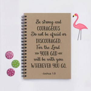 May include: A brown notebook with a spiral binding and a quote from Joshua 1:9. The quote reads "Be strong and courageous. Do not be afraid or discouraged. For the Lord your God will be with you wherever you go." The notebook is on a white surface with a pink flamingo and polka dot buttons.