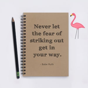May include: A brown notebook with a quote by Babe Ruth that reads "Never let the fear of striking out get in your way." The notebook has a spiral binding and is sitting on a white surface. A pink flamingo paper cutout is on the right side of the image.