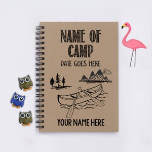 May include: A brown notebook with a spiral binding. The cover has a black and white illustration of a canoe on a lake with mountains in the background. The text "NAME OF CAMP" and "DATE GOES HERE" is printed on the cover. The text "YOUR NAME HERE" is printed at the bottom of the cover.