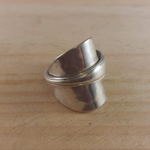 May include: A silver ring, crafted from repurposed silverware. The ring has a distinctive design with two curved sections overlapping. The ring is set against a light brown wooden surface.