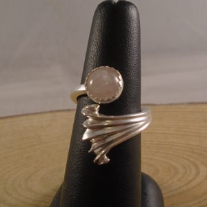 May include: A silver ring featuring a pink gemstone set in a bezel setting. The ring band is designed with a wave-like pattern.