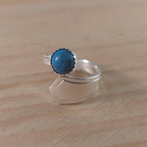 May include: A silver ring featuring a round turquoise stone set in a scalloped bezel. The ring's band is made from a repurposed spoon, with the spoon's handle forming the ring's shank. The turquoise stone is approximately 1.3 cm in diameter.