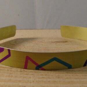 May include: A yellow metal headband with a colourful geometric pattern. The pattern includes purple, pink, teal, and orange shapes.