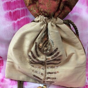 May include: A beige drawstring bag with a peacock feather design made of sequins and beads. The top of the bag is a darker brown fabric with gold accents. The drawstring is a twisted brown cord with a decorative bead.