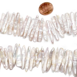 May include: Two strands of white, elongated freshwater pearls are displayed against a white background. A penny is placed near the pearls for size comparison. The pearls have a lustrous, iridescent sheen.
