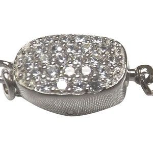 Peut inclure: Un fermoir de bijou argenté de forme rectangulaire et arrondie. La surface supérieure est recouverte de petites pierres claires et scintillantes. Le fermoir possède deux boucles pour l'attache aux bijoux.