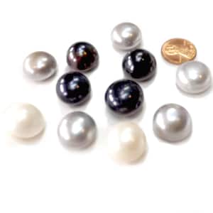 May include: A collection of lustrous pearls in various colors, including white, gray, and black, are scattered on a white surface. A copper penny is included for size comparison. The pearls have a smooth, rounded shape.