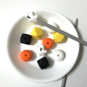May include: A white plate with a variety of silicone beads in orange, yellow, black, and white. The beads are shaped like pumpkins, ghosts, and diamonds. There are two gray knitting needles on the plate.