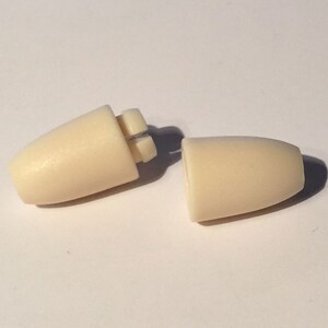 Puede incluir: Dos tapones para los o&iacute;dos de pl&aacute;stico de color blanquecino, uno con una costura visible, se muestran sobre una superficie blanca.