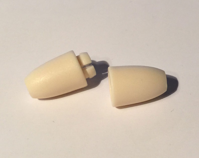 Puede incluir: Dos tapones para los o&iacute;dos de pl&aacute;stico de color blanquecino, uno con una costura visible, se muestran sobre una superficie blanca.
