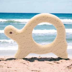 May include: A wooden turtle toy with a ring in the center. The toy is light brown and has a smooth finish. The turtle is on a sandy beach with blue ocean waves in the background.