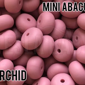 May include: A collection of matte orchid pink mini abacus beads, each featuring a central hole. The text MINI ABACUS is displayed at the top, and the word ORCHID is printed at the bottom, with the brand name TESLABABY visible in the background.