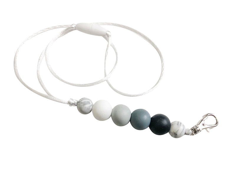 Puede incluir: Un cord&oacute;n blanco con un clip plateado y una cadena de cuentas de silicona en tonos grises y blancos. Las cuentas tienen un efecto marmoleado.