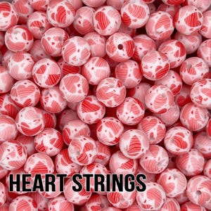 May include: A collection of spherical beads featuring a white base with a repeating red heart pattern. Each bead has a central hole for threading. The text HEART STRINGS is printed in bold black capital letters at the bottom of the image.