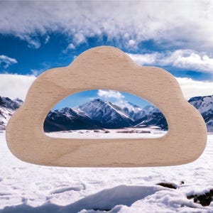 May include: A light-colored wooden cutout shaped like a cloud with a large, open, arched center. The smooth, natural wood grain is visible across the surface of the object, which frames a view of a snowy mountain landscape under a blue sky.
