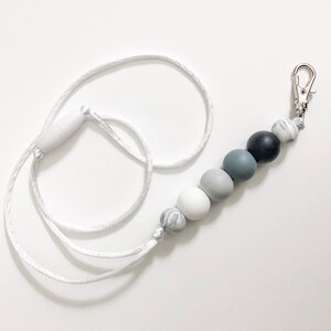 Puede incluir: Un cord&oacute;n blanco con un clip plateado y una cadena de cuentas de silicona grises, blancas y negras.
