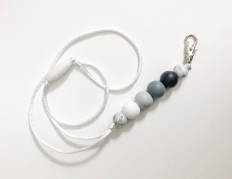 Puede incluir: Un cord&oacute;n blanco con un clip plateado y una cadena de cuentas de silicona grises, blancas y negras.