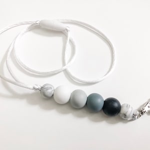 Puede incluir: Un cord&oacute;n blanco con un clip plateado y una cadena de cuentas de silicona grises y blancas. Las cuentas est&aacute;n en un patr&oacute;n de degradado, comenzando con blanco y terminando con gris.