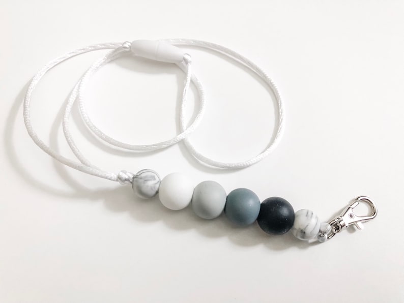 Puede incluir: Un cord&oacute;n blanco con un clip plateado y una cadena de cuentas de silicona grises y blancas. Las cuentas est&aacute;n en un patr&oacute;n de degradado, comenzando con blanco y terminando con gris.