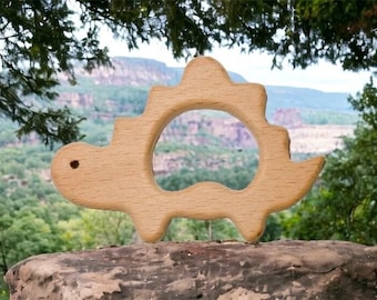 May include: A light brown wooden toy shaped like a dinosaur with a smooth, sanded finish. The figure features a rounded head with a small circular eye cutout, a jagged back profile, and a large central opening in the body for easy grasping.
