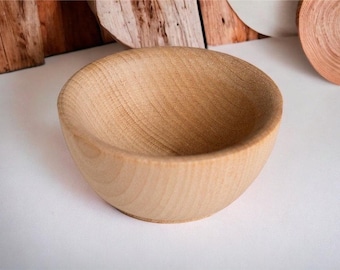 May include: A small, round, light-colored wooden bowl with a smooth, matte finish. The bowl features a visible natural wood grain pattern throughout its interior and exterior surface, showcasing a simple, curved design with a wide, open top.