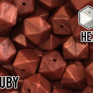 May include: A collection of metallic ruby-colored hexagonal beads with a central hole. The beads have a faceted geometric shape and a smooth, reflective finish. The text "RUBY" appears in the bottom left corner and "HEX" in the top right corner.