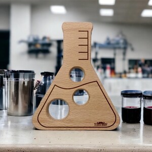 May include: A light-colored wooden Erlenmeyer flask-shaped board featuring three circular cutouts and etched measurement markings on the neck. The base of the wooden object displays the brand name TESLA BABY in dark lettering with a small mustache icon.