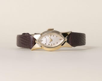 May include: A vintage wristwatch featuring a gold-tone marquise-shaped case with a white dial, gold hands, and small hour markers. The watch is attached to a dark brown leather strap with a textured, lizard-like embossed pattern and a gold crown.