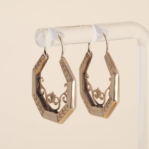 May include: A pair of gold-toned hoop earrings featuring an octagonal shape with engraved leaf patterns along the outer edges and intricate, delicate filigree scrollwork detailing suspended within the center of each hoop frame.