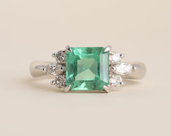 Könnte beinhalten: Ein silberfarbener Ring mit einem zentralen, quadratisch geschliffenen grünen Smaragd. Der Smaragd ist auf jeder Seite von einer Gruppe aus drei kleinen weißen Diamanten umgeben, darunter zwei Rundschliffe und ein Marquise-Schliff in Krappenfassung.