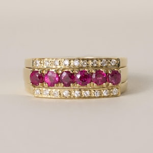 Puede incluir: Un anillo de oro con una fila central de seis rubíes redondos de color rojo rosado vibrante, flanqueados por dos filas exteriores de pequeños diamantes redondos transparentes. La banda tiene un acabado pulido con detalles milgrain en los bordes.