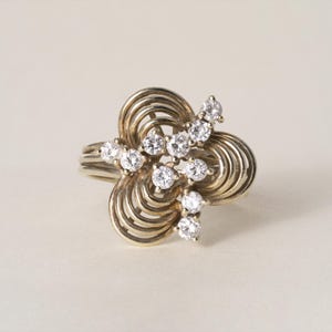 Könnte beinhalten: Ein goldfarbener Ring mit floralem Design. Der Ring hat eine Ansammlung kleiner, runder, klarer Edelsteine, die auf einer wirbelnden, durchbrochenen Basis angeordnet sind. Das Band ist schlicht und glatt.