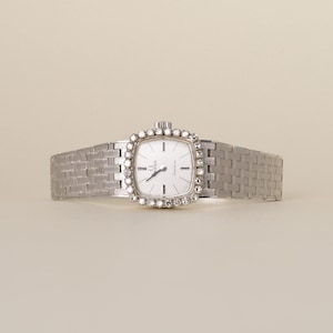May include: A silver-toned Omega Genève wristwatch featuring a square-shaped face with a white dial, silver hands, and hour markers. The bezel is surrounded by a border of clear stones, and the watch has a textured, woven-style metal link band.