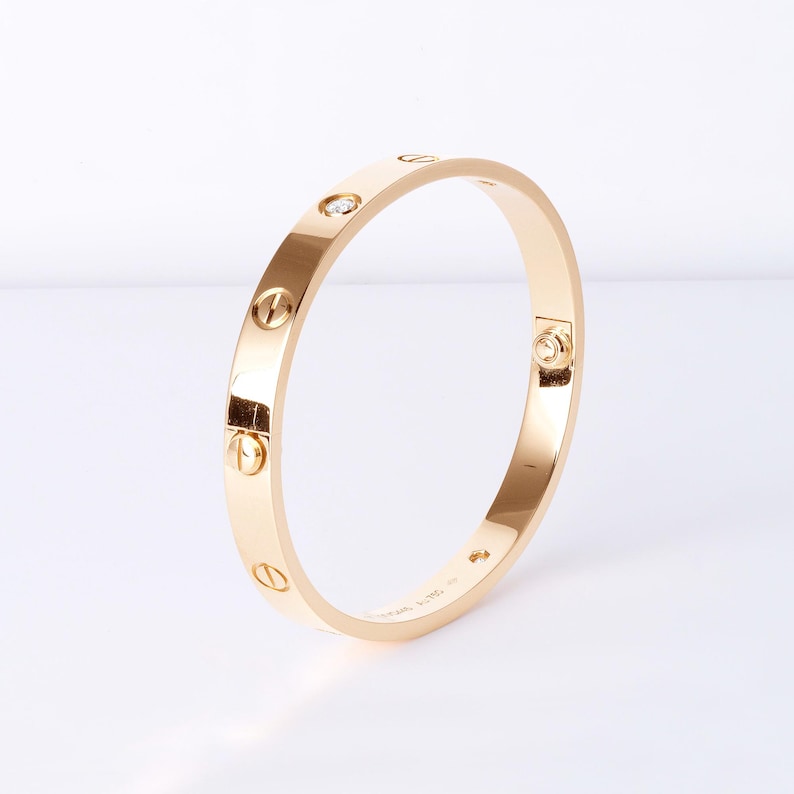 May include: A gold bangle bracelet with a diamond set in the center. The bracelet is decorated with screws around the edge. The bracelet is made by Cartier and has the inscription 'Cartier 750' on the inside.