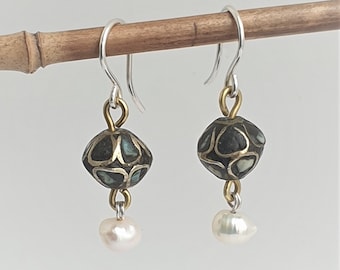 Op de afbeelding: Een paar oorhangers met zilveren haken, goudkleurige connectoren en donkere, bewerkte kralen. Elke oorbel heeft een kleine, witte, druppelvormige parel. De oorbellen worden gepresenteerd op een dunne, bruine houten stok.